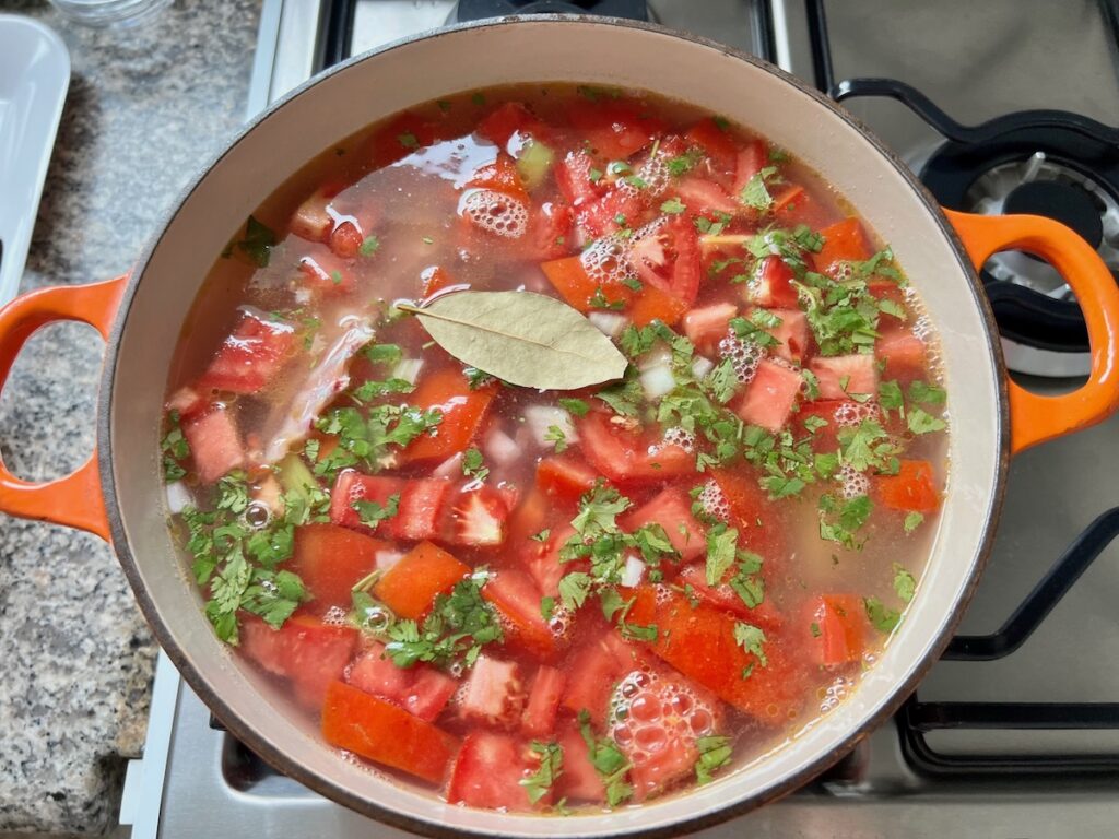 Water, bay leaf, minced garlic, chopped parsley, and chopped fresh tomatoes added to pot with raw chicken for the Canja Brazilian Chicken and Rice Soup recipe.