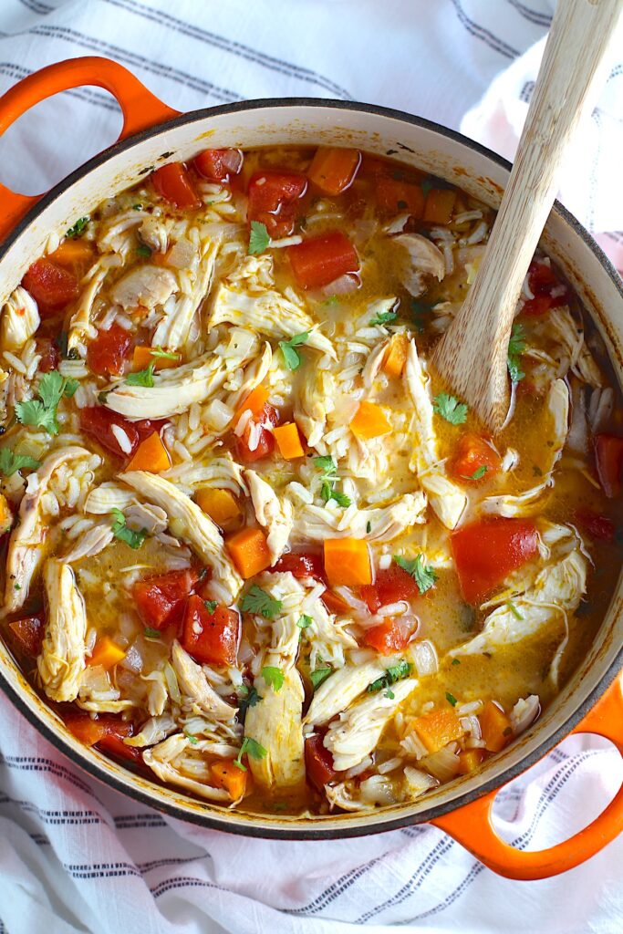 Canja Chicken Rice Soup in a pot with carrots and tomatoes and parsley and a wooden spoon on counter.