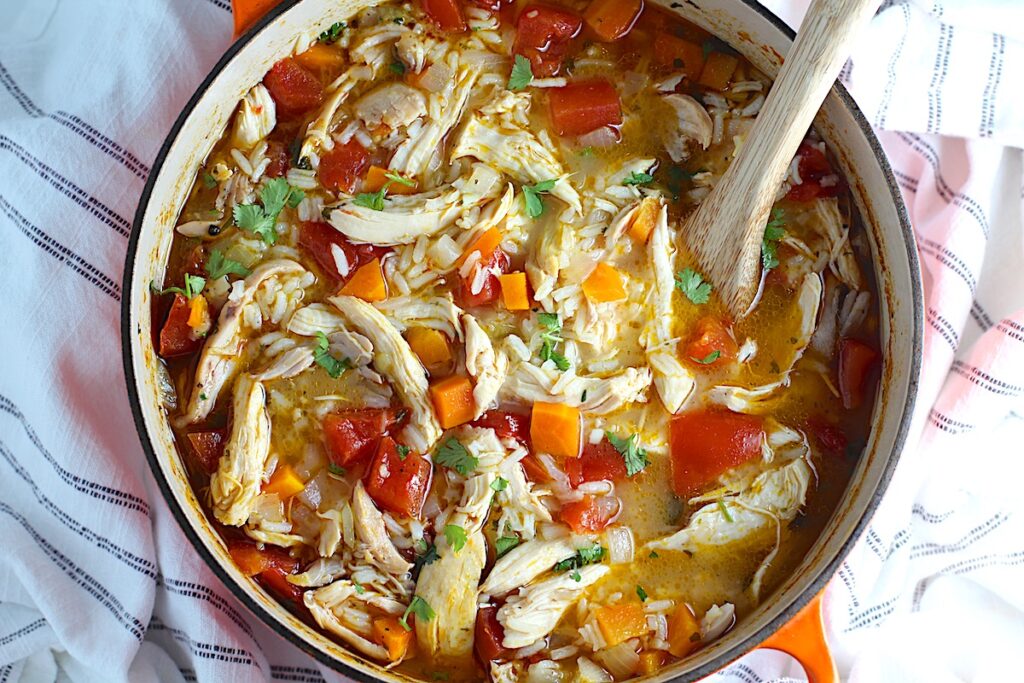 Canja Chicken Rice Soup in a pot with carrots and tomatoes and parsley and a wooden spoon on counter.