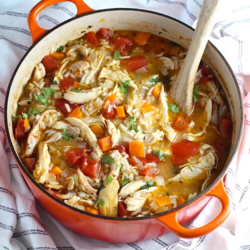 Canja Chicken Rice Soup in a pot with carrots and tomatoes and parsley and a wooden spoon on counter.