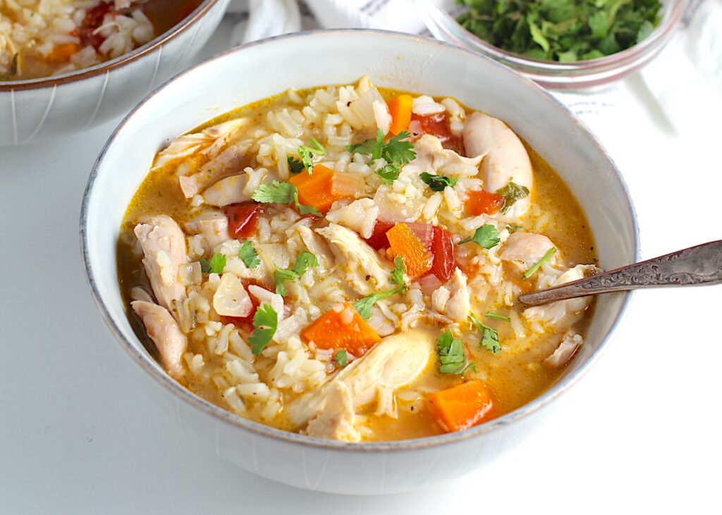 Canja Chicken Rice Soup in a bowl with carrots and tomatoes and parsley and a spoon on counter.
