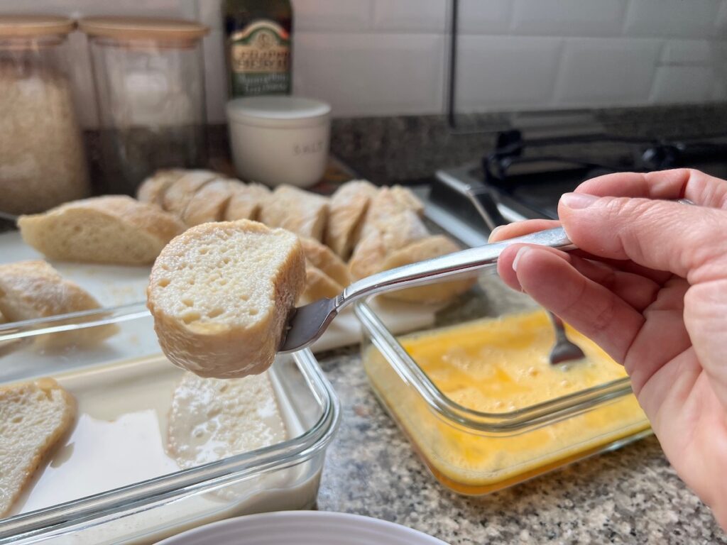 Hand with fork holding a baguette slice soaked in the milk mixture over the bowl to let excess drip off before placing it in the second dish with beaten eggs for Brazilian French Toast with Condensed Milk.