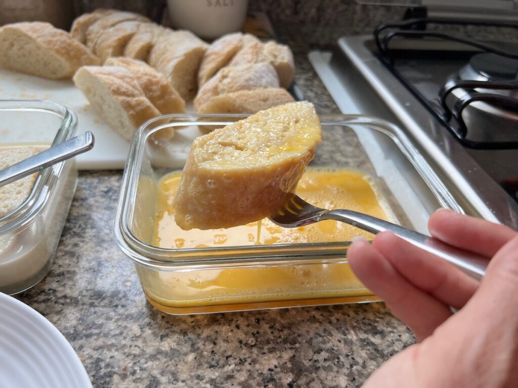 Hand with fork holding a baguette slice soaked in the egg mixture over the bowl to let excess drip off before placing it a dish to fry for Brazilian French Toast with Condensed Milk.