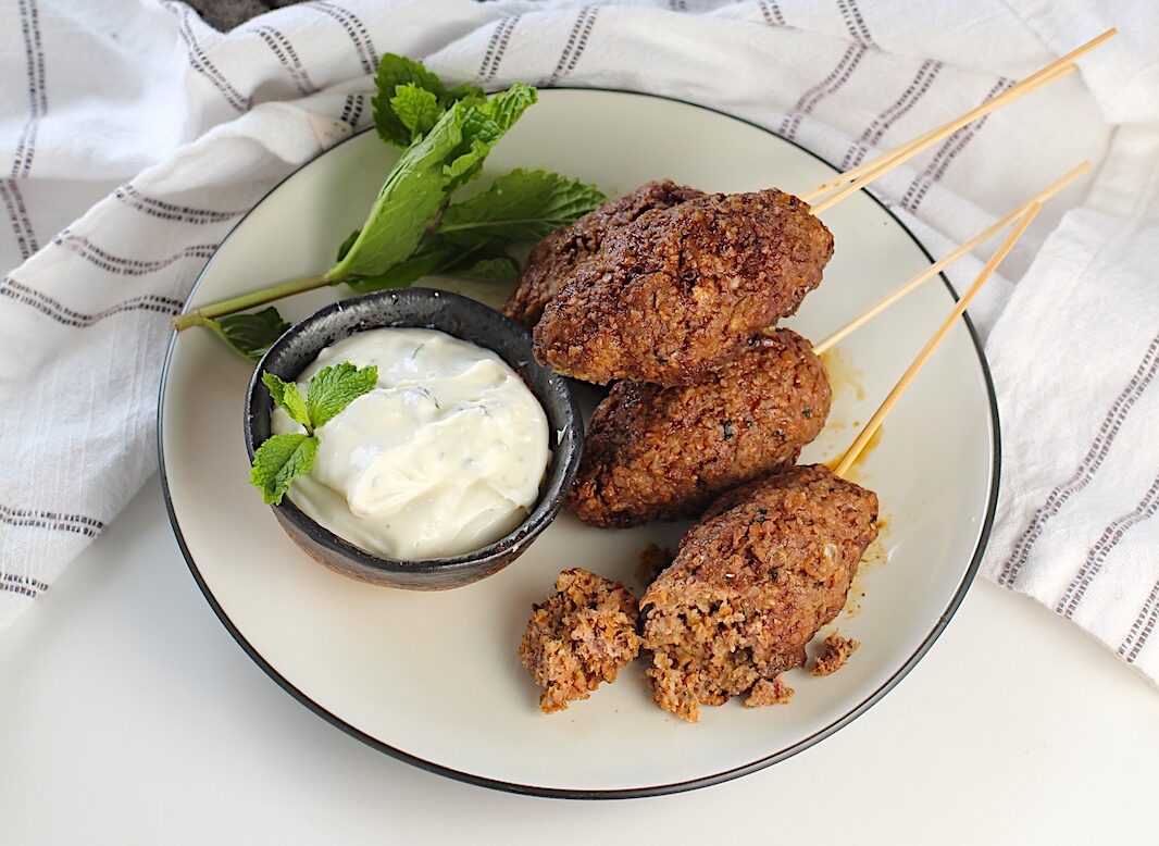 Four Kibe kabobs, Mediterranean Ground Beef Kabob recipe, cooked, skewered, and plated with yogurt dipping sauce and fresh mint leaves as garnish. One Kibe on the plate has been cut open to expose the tender meat mixture inside.