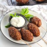 Four Kibe kabobs, Mediterranean Ground Beef Kabob recipe, cooked, skewered, and plated with yogurt dipping sauce and fresh mint leaves as garnish.