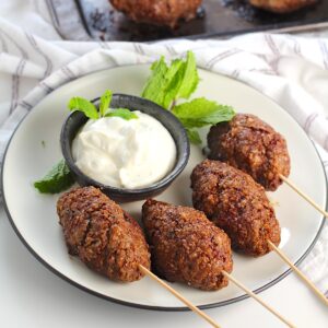 Four Kibe kabobs, Mediterranean Ground Beef Kabob recipe, cooked, skewered, and plated with yogurt dipping sauce and fresh mint leaves as garnish.