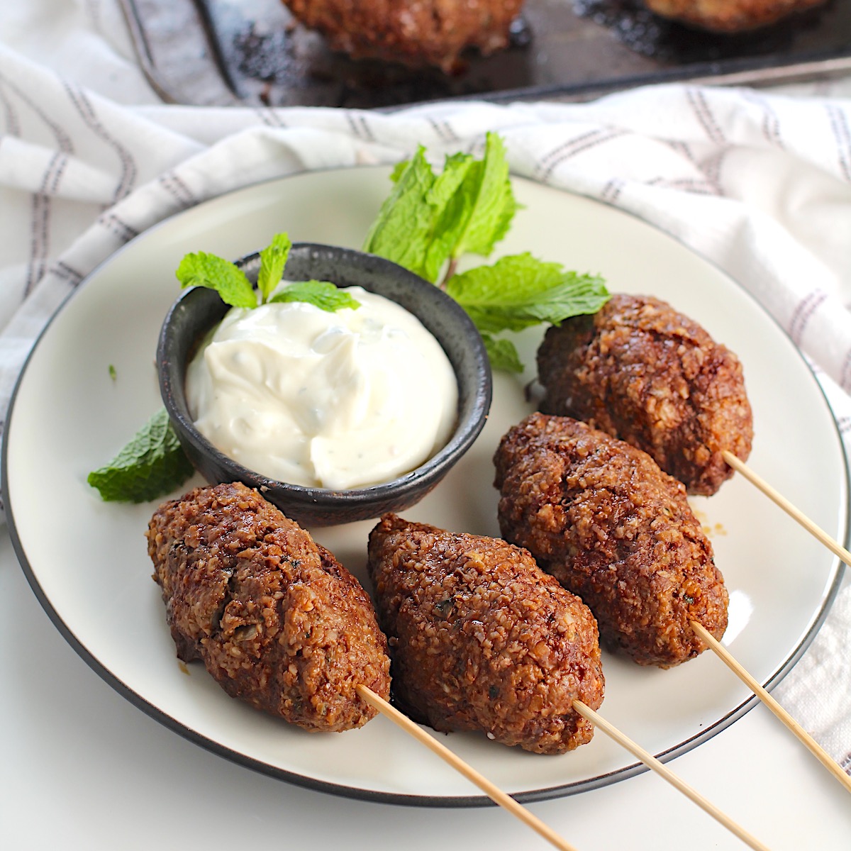 Four Kibe kabobs, Mediterranean Ground Beef Kabob recipe, cooked, skewered, and plated with yogurt dipping sauce and fresh mint leaves as garnish.
