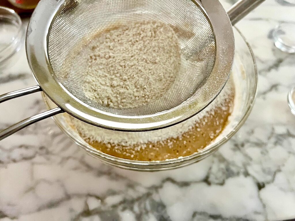 Flour being sifted into batter in a clear glass bowl before being mixed for the Pao de Mel recipe, Brazilian cakes filled with dolce de leite and covered with chocolate.