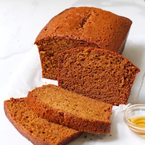 Honey Loaf cake baked beautifully golden brown on table with 3 slices standing in front showing the moist inside. On the counter is a small bowl of honey.