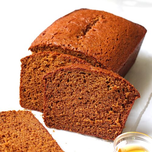 Honey Loaf cake baked beautifully golden brown on table with 3 slices standing in front showing the moist inside. On the counter is a small bowl of honey.
