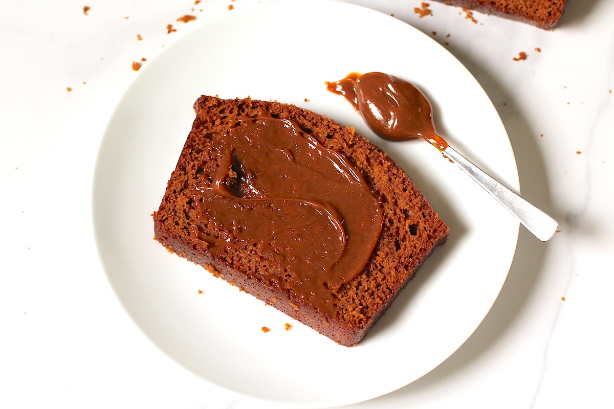Slice of Honey Loaf cake on a plate with doce de leite spread on top and a spoon on the plate with more doce de leite.baked beautifully golden brown on table with 3 slices standing in front showing the moist inside. On the counter is a small bowl of honey.