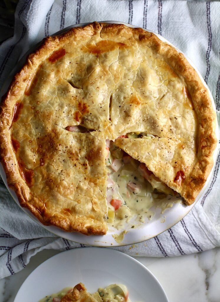 Ham and Leek Pie with potatoes and carrots in a cream sauce and flaky golden brown pie crust on top.  Pie in pie pan on counter with 1 slice removed showing creamy, savory filling.