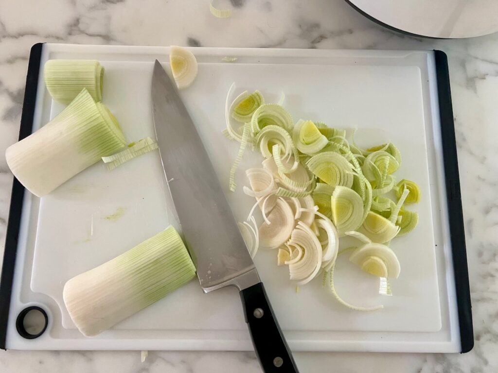 Raw white ends of leeks being sliced thin with a chef's knife on cutting board for the Ham and Leek Pie.