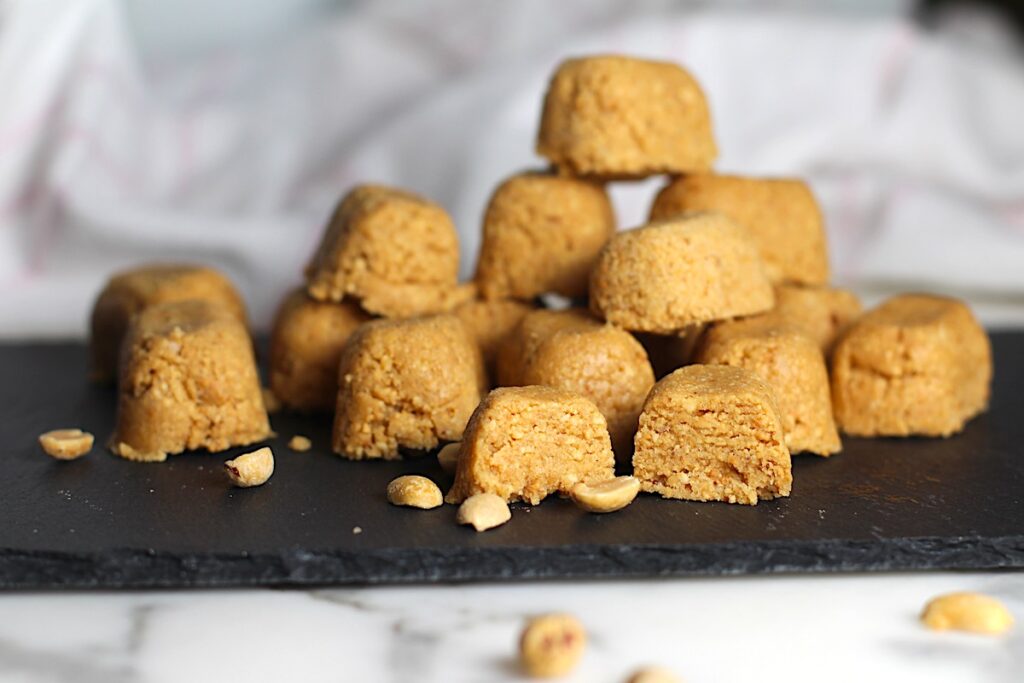 Twelve small mounds of Brazilian Peanut Candy or This Paçoca de Amendoim, stacked on a black platter with one cut open in the front, peanut halves on the platter and table, and a towel in the background.