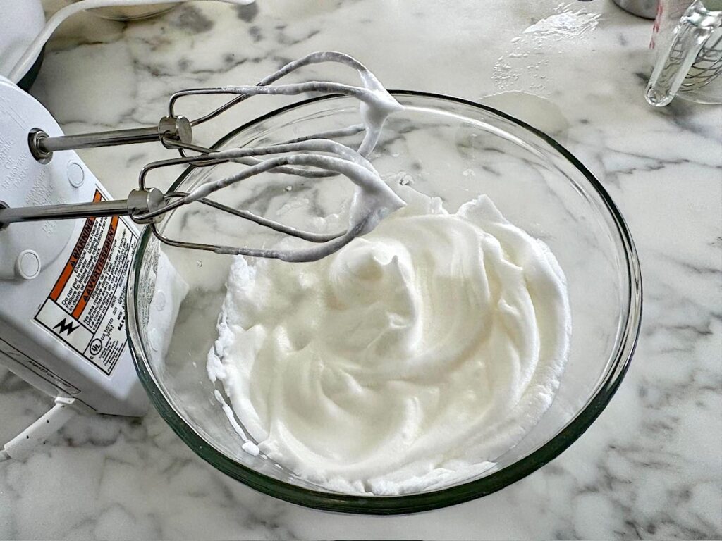 Egg whites whipped in a glass bowl with hand mixer showing soft peaks for this Bolo de Fuba, Cornmeal Cake, recipe.
