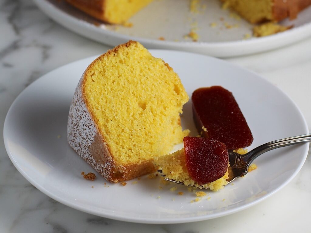 Piece of Bolo de Fuba (cornmeal Bundt Cake) on plate with fork holding a bite with guava paste on top and on the side. The whole cake with missing piece is in background.