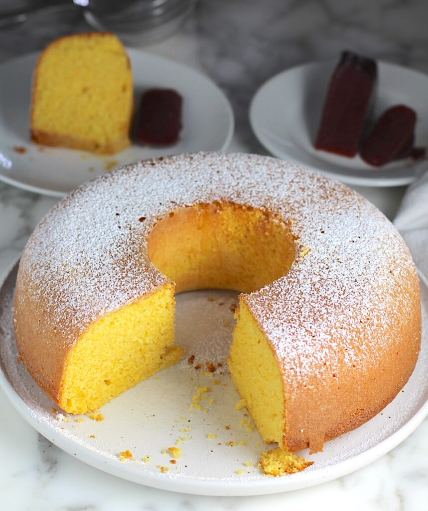 Bolo de Fuba (cornmeal Bundt Cake) on plate with powdered sugar dusted on top and large piece removed from the front of the bundt cake. Slice of cake on a plate in the back and guava paste on another plate in the back.