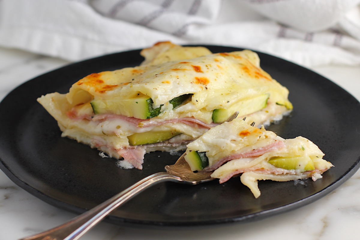 Serving of Bechamel Sauce Lasagna on a black plate with fork that has cut through the front to show the layers of cheese, zucchini, ham, and pasta. 