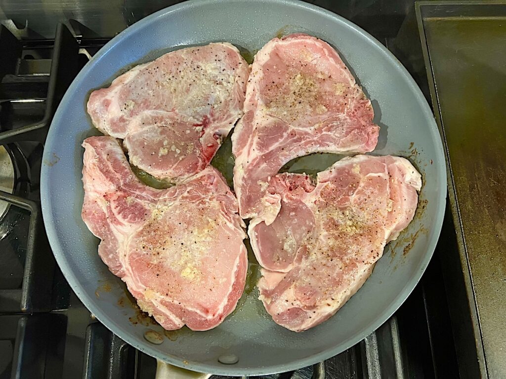 Raw marinated bone-in pork chops added to frying pan for this Pork Chops with Onions recipe.