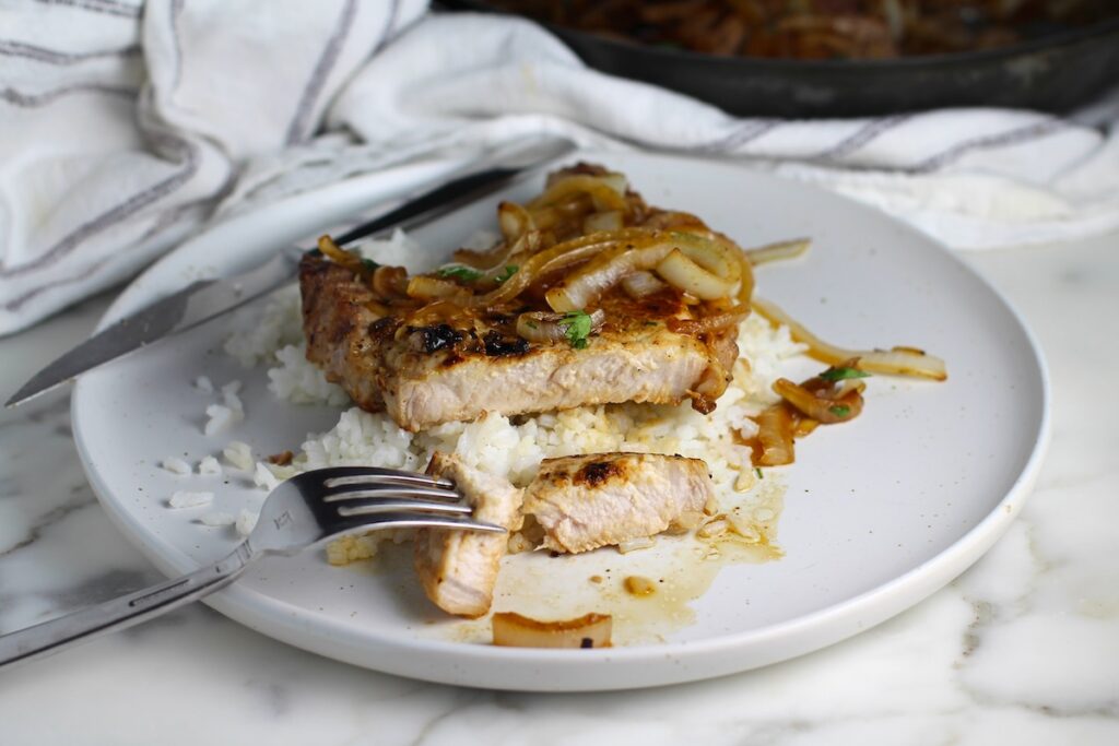 Browned cooked bone-in pork chop Sliced with fork holding a bite. The pork is topped with sauteed onions all over white rice on a plate for this Pork Chops with Onions recipe, Costeletas de Porco com Cebolas.