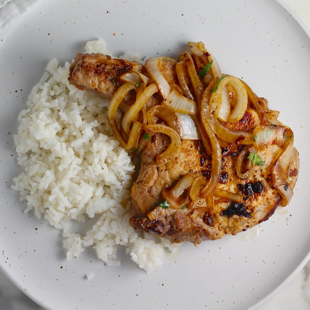 Browned cooked bone-in pork chop topped with sauteed onions all over white rice on a plate for this Pork Chops with Onions recipe, Costeletas de Porco com Cebolas.