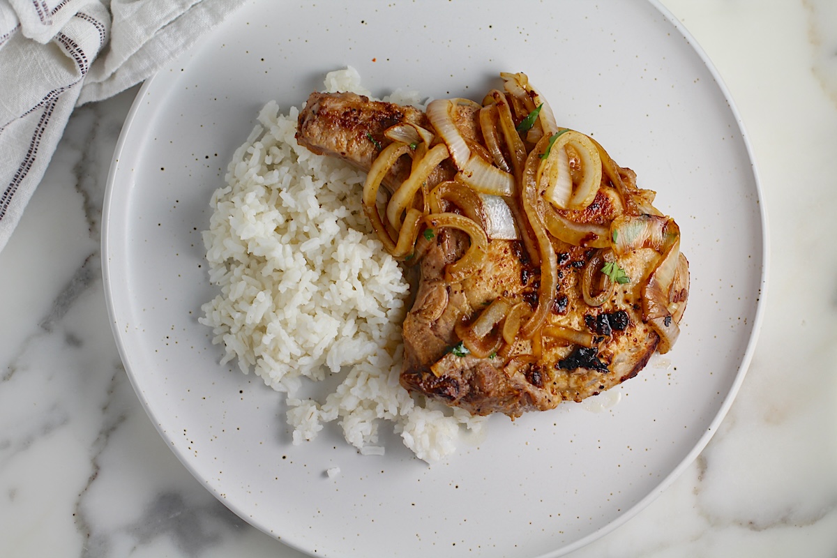 Browned cooked bone-in pork chop topped with sauteed onions all over white rice on a plate for this Pork Chops with Onions recipe, Costeletas de Porco com Cebolas.