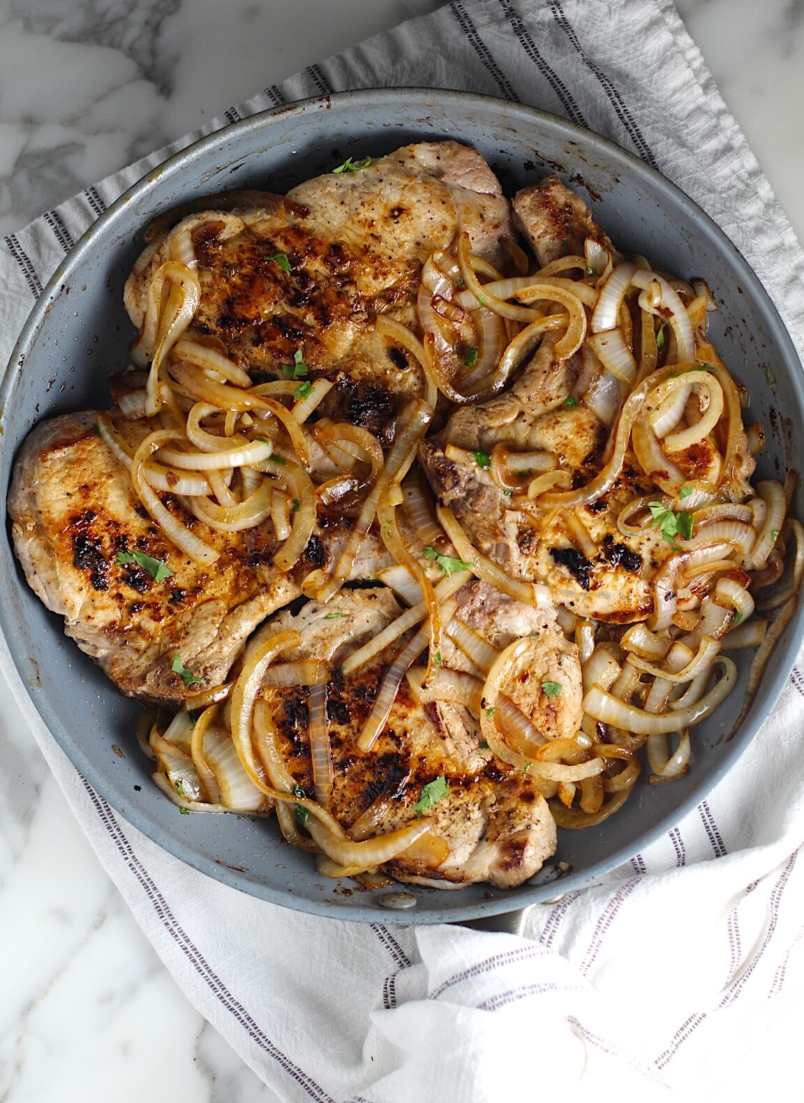 Browned cooked sliced onions topped on cooked bone-in pork chops in frying pan for this Pork Chops with Onions recipe, Costeletas de Porco com Cebolas.