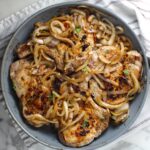 Browned cooked sliced onions topped on cooked bone-in pork chops in frying pan for this Pork Chops with Onions recipe, Costeletas de Porco com Cebolas.