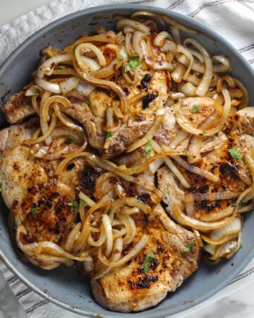 Browned cooked sliced onions topped on cooked bone-in pork chops in frying pan for this Pork Chops with Onions recipe, Costeletas de Porco com Cebolas.