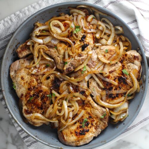 Browned cooked sliced onions topped on cooked bone-in pork chops in frying pan for this Pork Chops with Onions recipe, Costeletas de Porco com Cebolas.