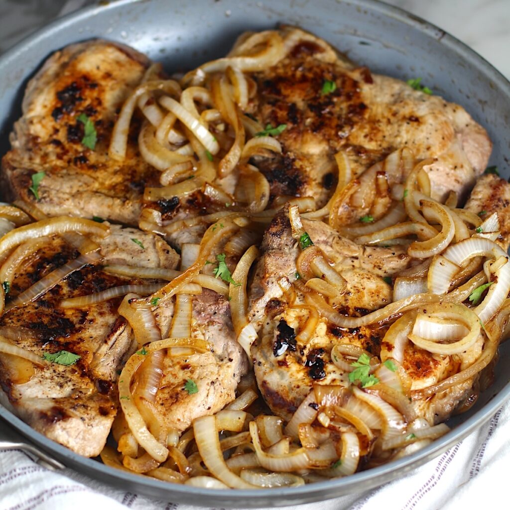 Browned cooked sliced onions topped on cooked bone-in pork chops in frying pan for this Pork Chops with Onions recipe, Costeletas de Porco com Cebolas.